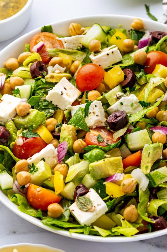 Close-up side view of Mediterranean chopped salad with romaine lettuce, chickpeas, cubed feta cheese, avocado, cucumber, cherry tomatoes, yellow bell pepper, red onion, Kalamata olives, parsley, and black pepper in a white bowl.