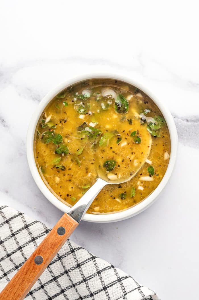 Overhead view of lemon-Dijon dressing mixed in a small white bowl with a spoon, showing olive oil, herbs, garlic, black pepper, and lemon juice on a white marble surface beside a black-and-white checkered towel.