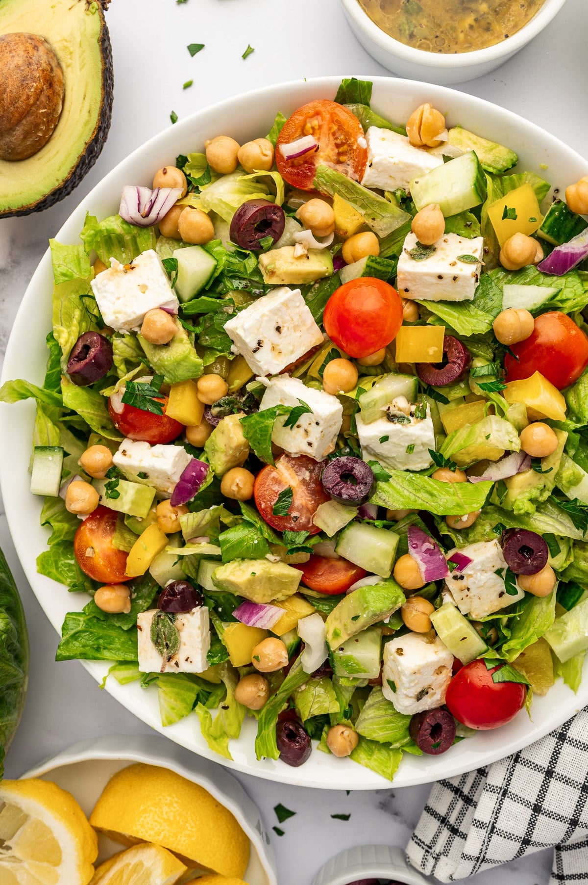 Overhead close-up of a Mediterranean chopped salad in a white bowl with romaine lettuce, chickpeas, cucumber, cherry tomatoes, red onion, yellow bell pepper, sliced Kalamata olives, cubed feta cheese, avocado, parsley, and black pepper, with lemon wedges, a small bowl of dressing, and a black-and-white checkered towel around the bowl.