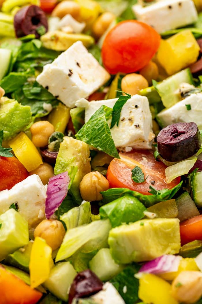 Extreme close-up of Mediterranean salad with cubed feta cheese, sliced Kalamata olives, chickpeas, avocado, cucumber, cherry tomatoes, yellow bell pepper, red onion, chopped parsley, and romaine lettuce.