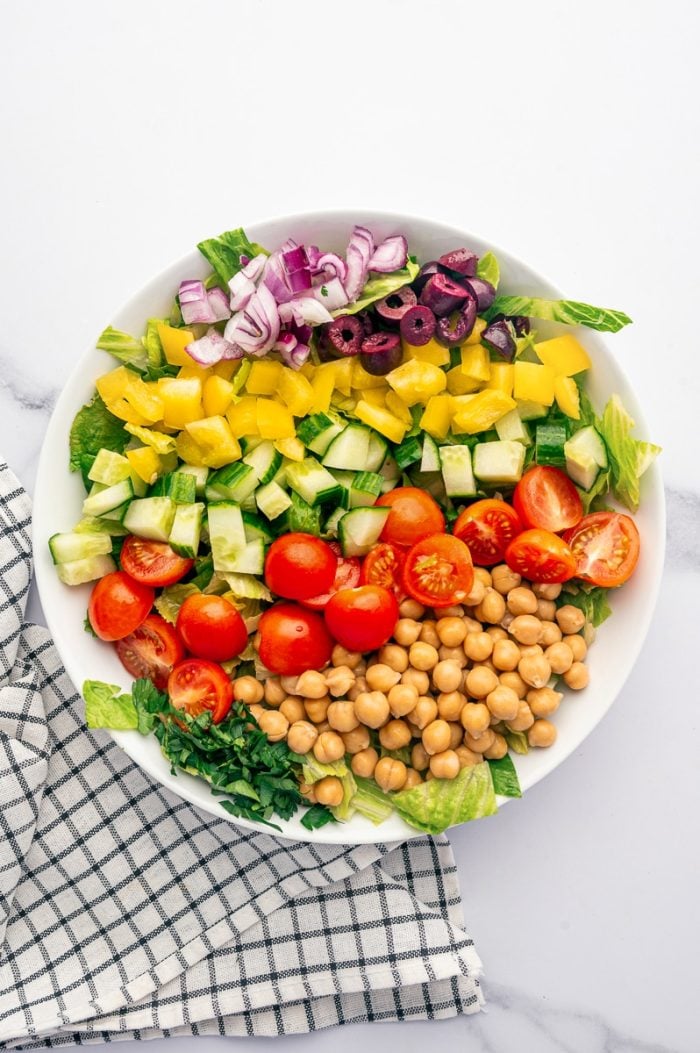 Overhead view of chopped Mediterranean salad ingredients arranged in a large white bowl, including romaine lettuce, chickpeas, diced cucumber, halved cherry tomatoes, yellow bell pepper, red onion, sliced Kalamata olives, and chopped parsley beside a black-and-white checkered towel.