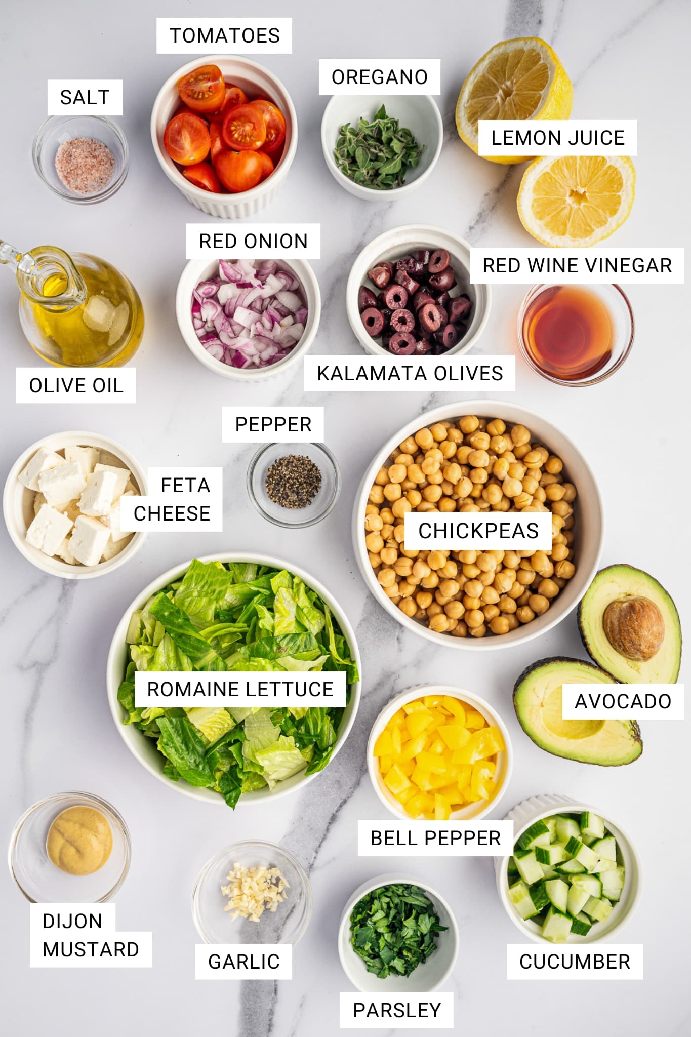 Overhead labeled ingredient photo for Mediterranean chopped salad on a white marble surface with text overlays for salt, tomatoes, oregano, lemon juice, red onion, red wine vinegar, Kalamata olives, olive oil, feta cheese, pepper, chickpeas, romaine lettuce, avocado, Dijon mustard, garlic, bell pepper, parsley, and cucumber.