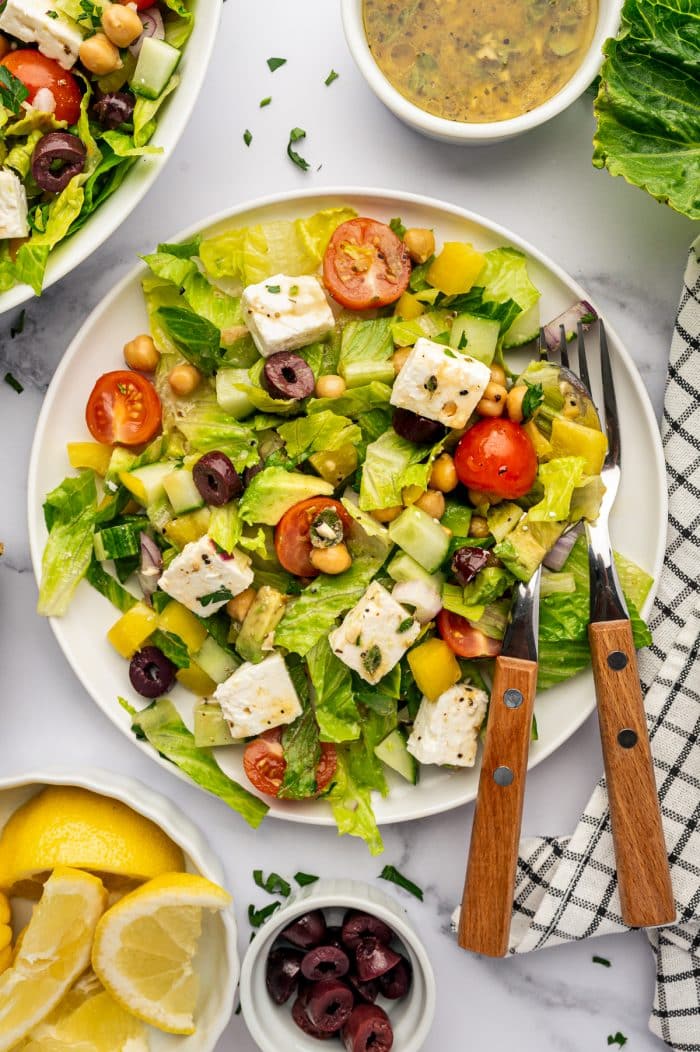 Overhead view of Mediterranean salad served on a white plate with romaine lettuce, chickpeas, cucumber, cherry tomatoes, yellow bell pepper, red onion, Kalamata olives, cubed feta cheese, avocado, and parsley, with wooden-handled salad servers, lemon wedges, extra olives, dressing, and a black-and-white checkered towel on a white marble surface.