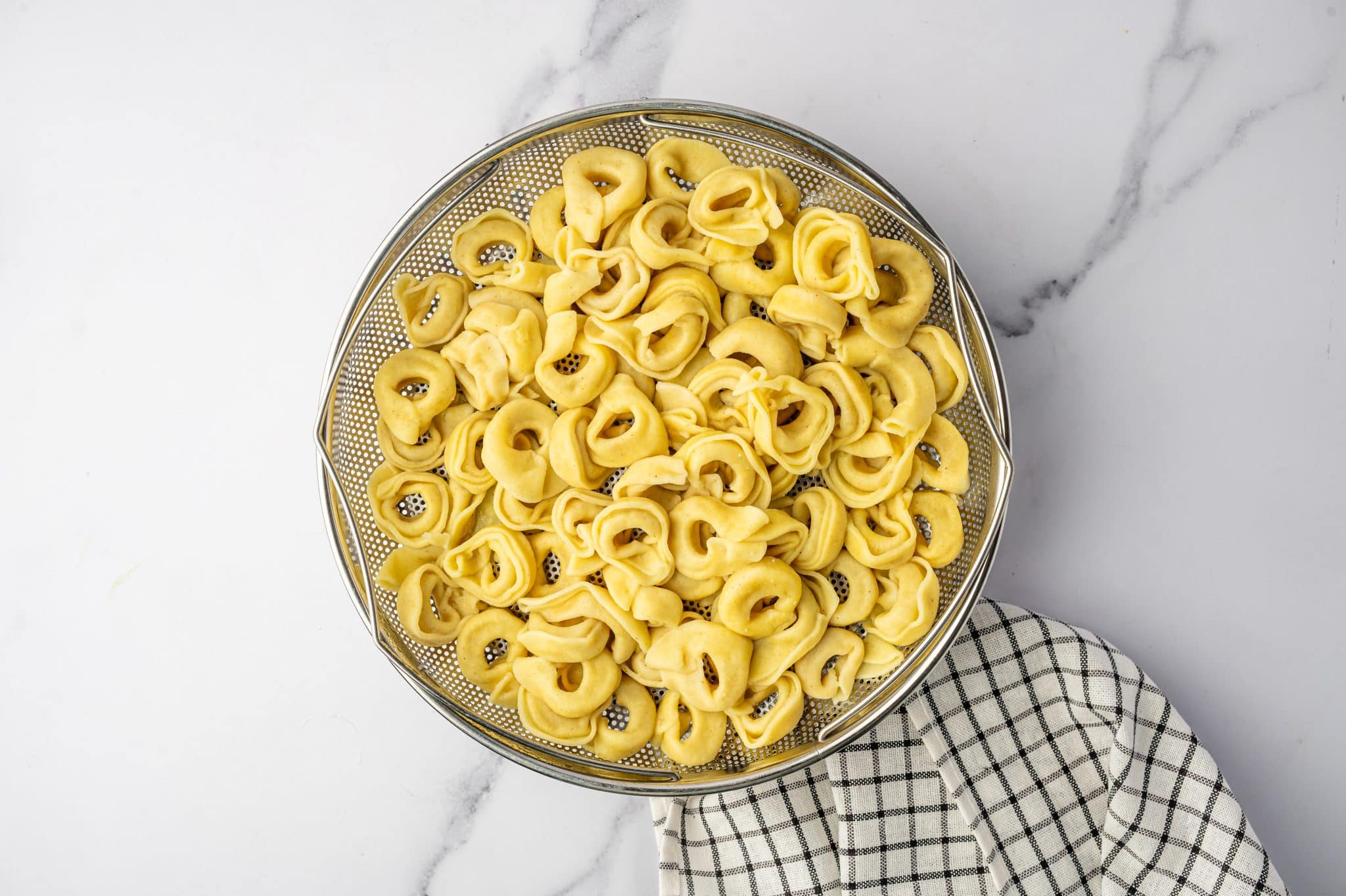 Overhead image of cooked cheese tortellini draining in a round metal colander on a white marble surface. The tortellini is pale yellow and piled in the center of the colander, with a black and white checkered kitchen towel partially visible along the lower right side.