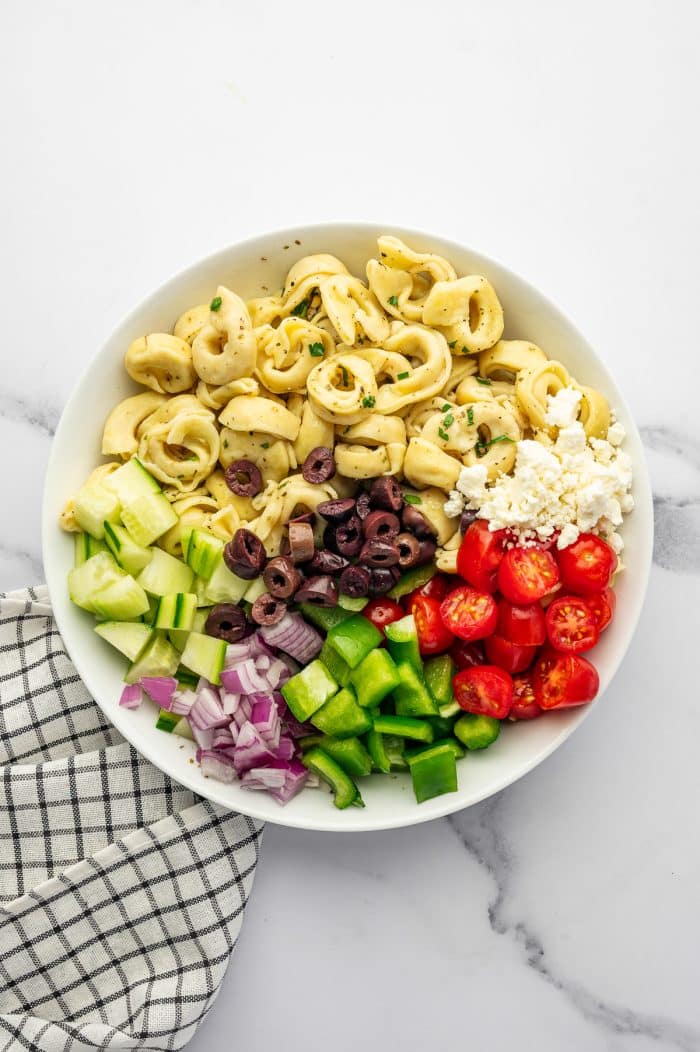 Overhead image of tortellini pasta salad ingredients arranged in a large white bowl on a white marble surface. The bowl contains cooked cheese tortellini, chopped cucumber, diced red onion, green bell pepper, halved cherry tomatoes, sliced kalamata olives, and crumbled feta cheese. A black and white checkered towel is visible along the left side.