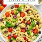 Pinterest pin image of tortellini pasta salad in a large white bowl with cheese tortellini, cherry tomatoes, cucumber, green bell pepper, red onion, kalamata olives, crumbled feta cheese, and fresh basil. A white text box overlay at the top reads "TORTELLINI PASTA SALAD" with "THEFORKEDSPOON.COM" underneath, and a small bowl of dressing is partially visible in the upper right.