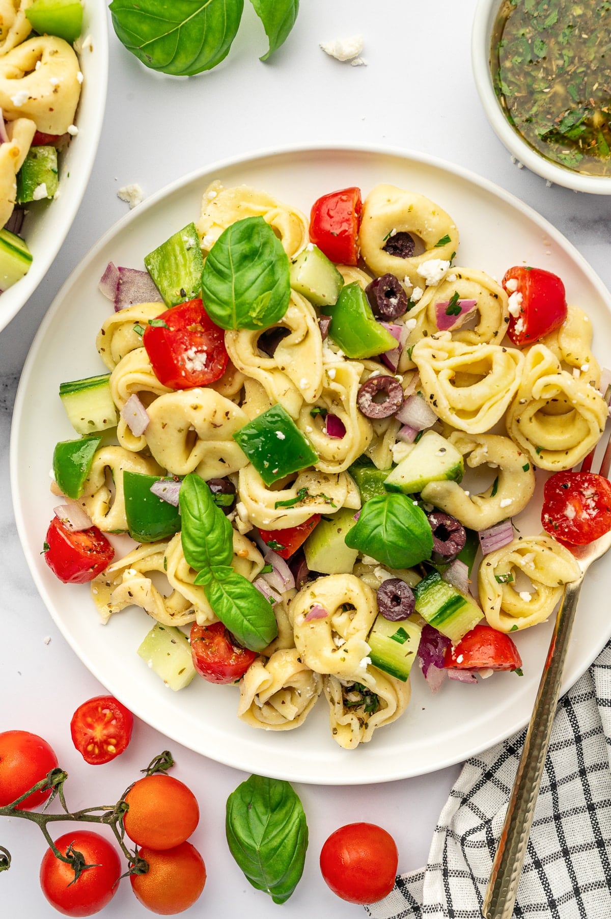 Overhead image of tortellini pasta salad served on a white plate with cheese tortellini, cucumber, green bell pepper, cherry tomatoes, red onion, kalamata olives, crumbled feta, and fresh basil leaves. A bowl of dressing is partially visible in the upper right, with vine tomatoes, loose basil leaves, and a black and white checkered towel around the plate.