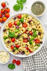 Overhead image of tortellini pasta salad in a large white bowl with cheese tortellini, cherry tomatoes, cucumber, green bell pepper, red onion, kalamata olives, crumbled feta, and fresh basil leaves. A small white bowl of homemade Italian dressing sits near the top right, with vine tomatoes, basil, extra feta, and a black and white checkered towel arranged around the salad.