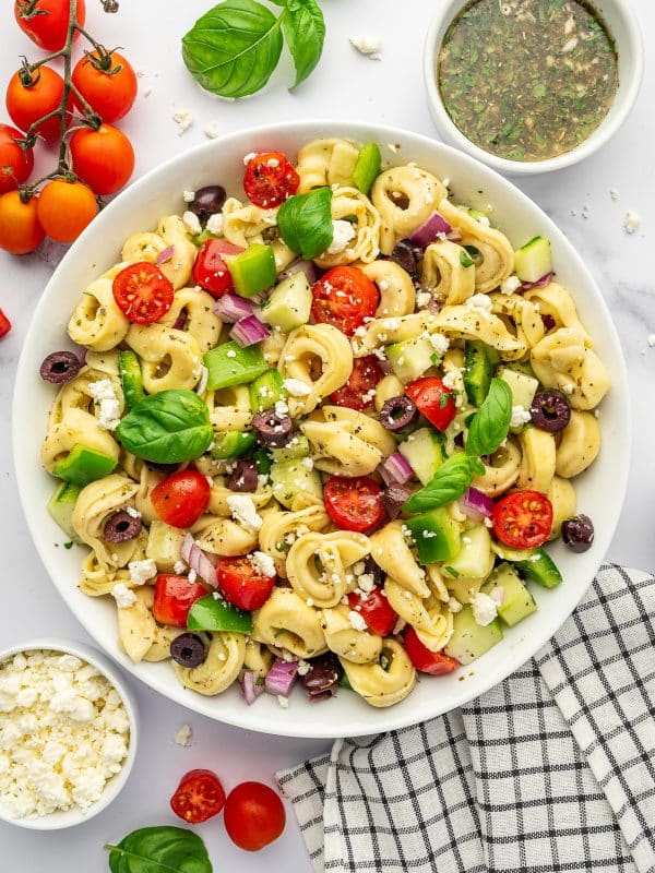 Overhead image of tortellini pasta salad in a large white bowl with cheese tortellini, cherry tomatoes, cucumber, green bell pepper, red onion, kalamata olives, crumbled feta, and fresh basil leaves. A small white bowl of homemade Italian dressing sits near the top right, with vine tomatoes, basil, extra feta, and a black and white checkered towel arranged around the salad.