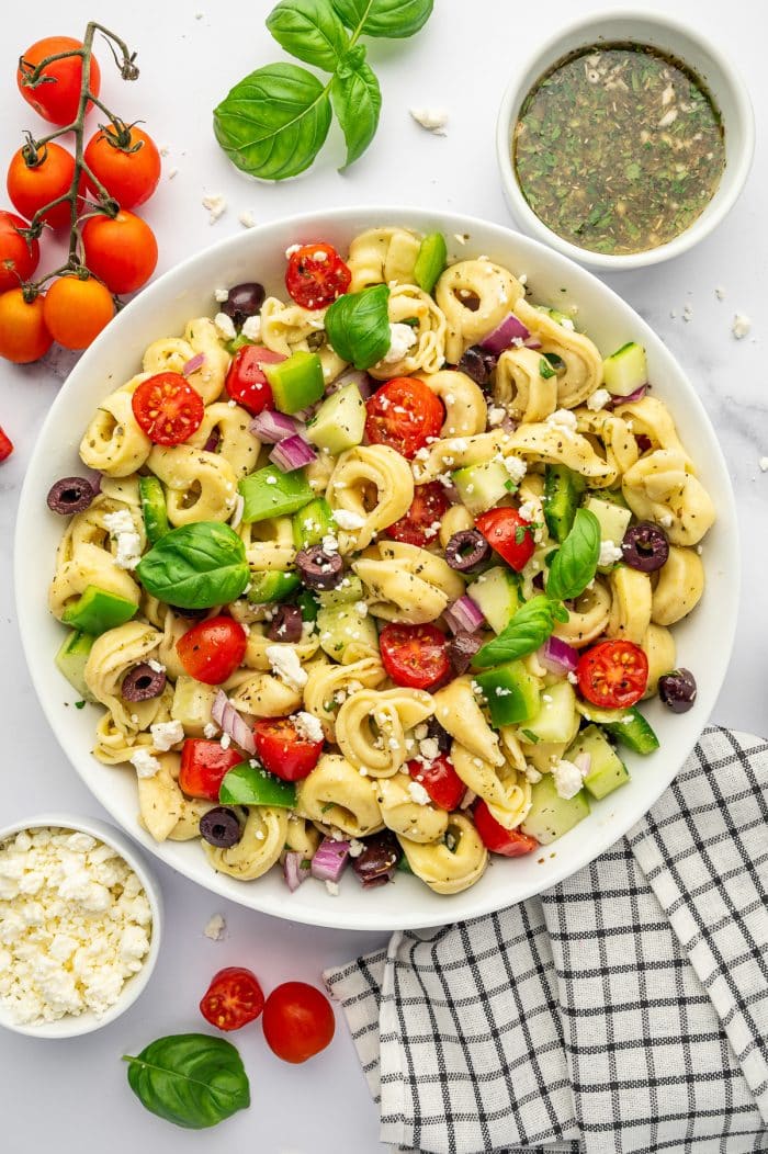 Overhead image of tortellini pasta salad in a large white bowl with cheese tortellini, cherry tomatoes, cucumber, green bell pepper, red onion, kalamata olives, crumbled feta, and fresh basil leaves. A small white bowl of homemade Italian dressing sits near the top right, with vine tomatoes, basil, extra feta, and a black and white checkered towel arranged around the salad.