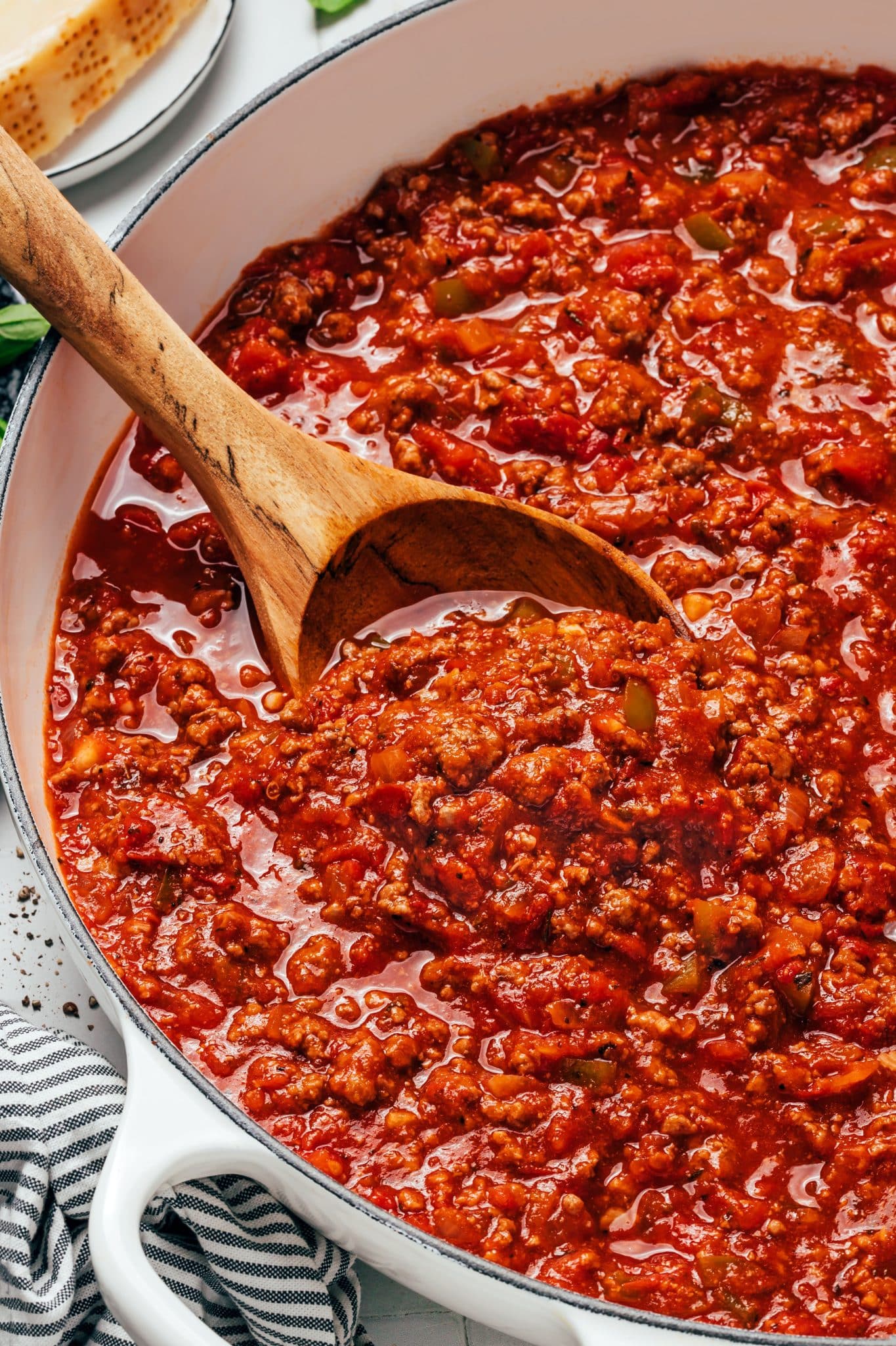 A large wooden serving spoon holds a generous scoop of thick red spaghetti sauce above a white enameled Dutch oven. The spoonful contains browned ground beef crumbles, softened pieces of onion and green bell pepper, and chunks of tomato coated in the rich sauce. More of the chunky meat sauce fills the pot below.