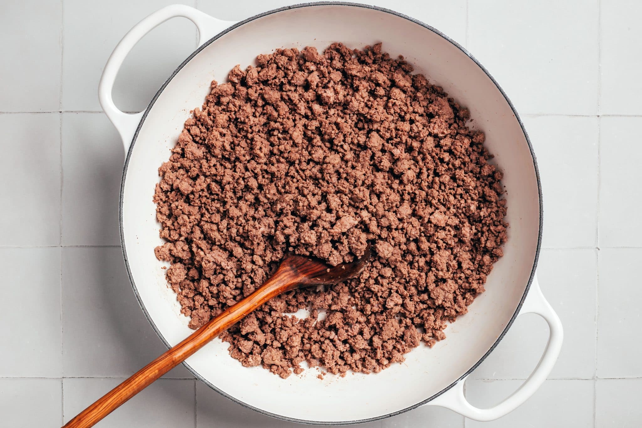 A large white enameled Dutch oven filled with browned ground beef broken into small, uneven crumbles. A long wooden cooking spoon rests among the meat, which has a light brown color with slightly darker browned pieces throughout.