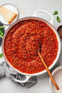 A large white enameled Dutch oven filled with thick, red ground beef spaghetti sauce. A long wooden serving spoon rests in the sauce, surrounded by visible pieces of ground beef, diced tomatoes, onions, and green bell peppers. A wedge of Parmesan cheese, fresh basil leaves, and a striped kitchen towel surround the pot.
