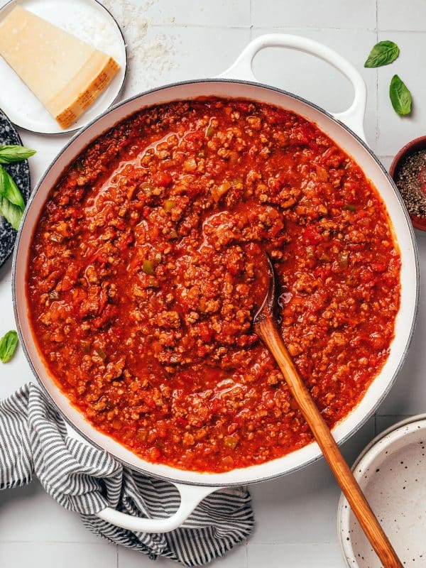 A large white enameled Dutch oven filled with thick, red ground beef spaghetti sauce. A long wooden serving spoon rests in the sauce, surrounded by visible pieces of ground beef, diced tomatoes, onions, and green bell peppers. A wedge of Parmesan cheese, fresh basil leaves, and a striped kitchen towel surround the pot.