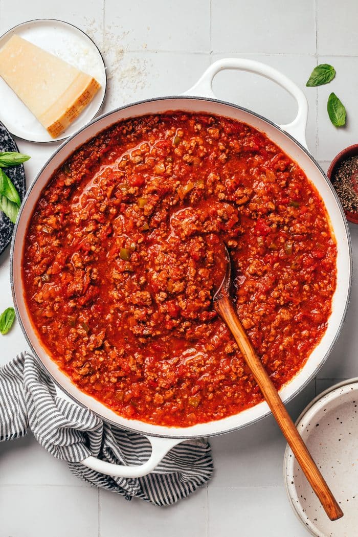 A large white enameled Dutch oven filled with thick, red ground beef spaghetti sauce. A long wooden serving spoon rests in the sauce, surrounded by visible pieces of ground beef, diced tomatoes, onions, and green bell peppers. A wedge of Parmesan cheese, fresh basil leaves, and a striped kitchen towel surround the pot.