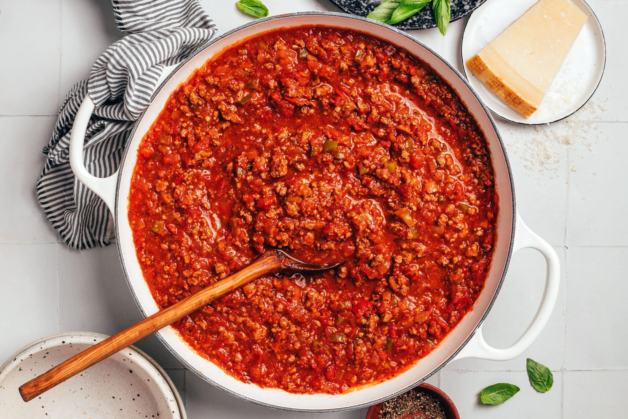 A large white enameled Dutch oven filled with thick, red ground beef spaghetti sauce. A long wooden serving spoon rests in the sauce, surrounded by visible pieces of ground beef, diced tomatoes, onions, and green bell peppers. A wedge of Parmesan cheese, fresh basil leaves, and a striped kitchen towel surround the pot.