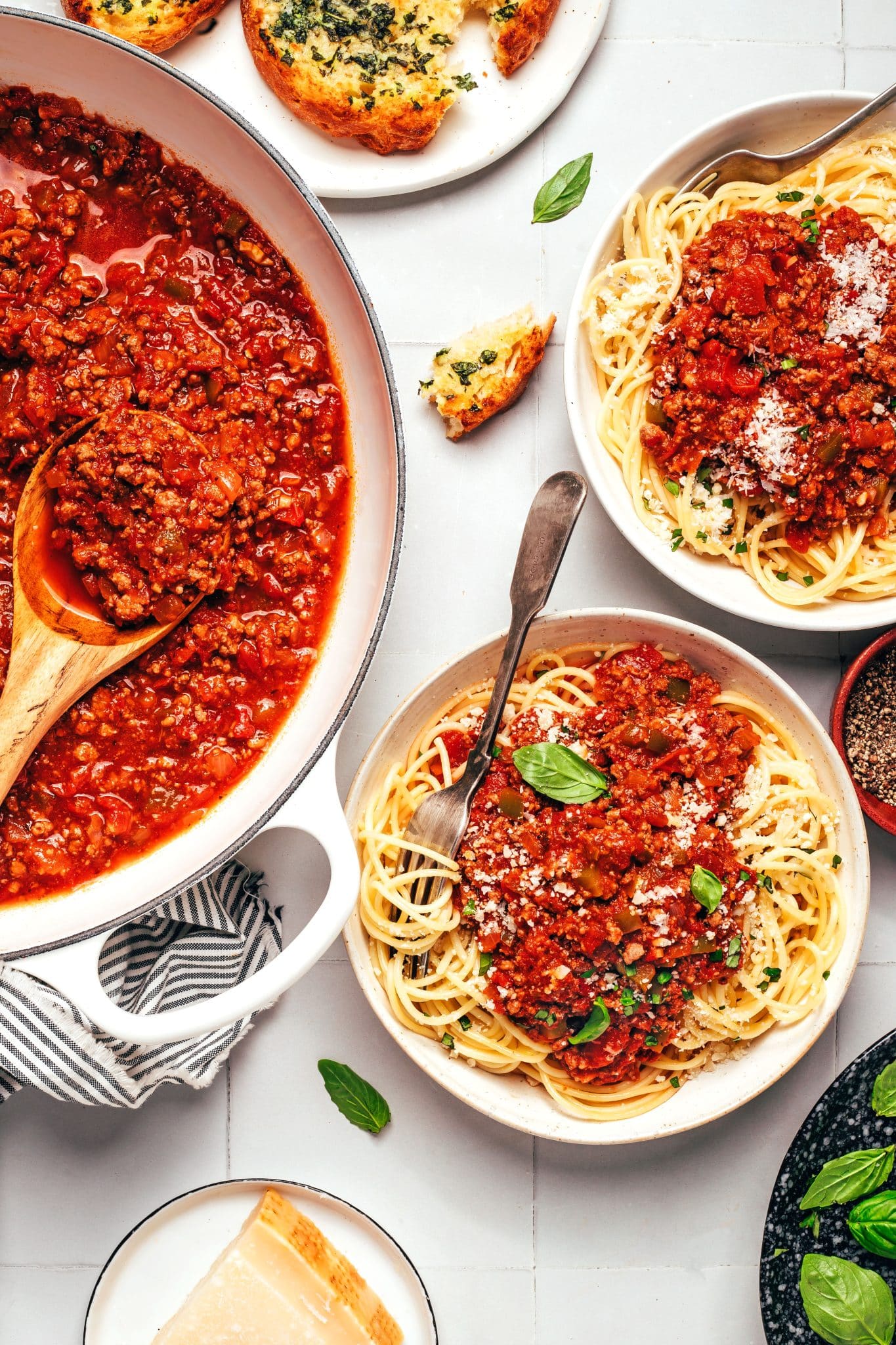 A large white enameled Dutch oven filled with chunky red ground beef spaghetti sauce sits beside two white bowls of spaghetti topped with the same meat sauce, grated Parmesan cheese, and fresh basil. A wooden spoon rests in the pot, while toasted garlic bread, a wedge of Parmesan cheese, scattered basil leaves, and a striped kitchen towel complete the dinner spread.