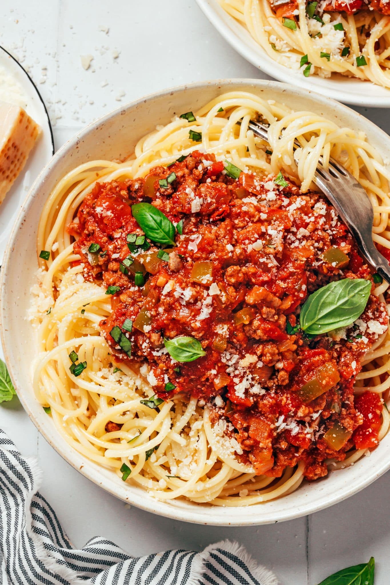 A white ceramic bowl filled with long spaghetti noodles topped with a thick, chunky red tomato and ground beef sauce. Pieces of diced green bell pepper, softened onions, and tomatoes are visible throughout the sauce. Fresh green basil leaves, chopped basil, and grated Parmesan cheese cover the pasta, with a metal fork resting among the noodles.