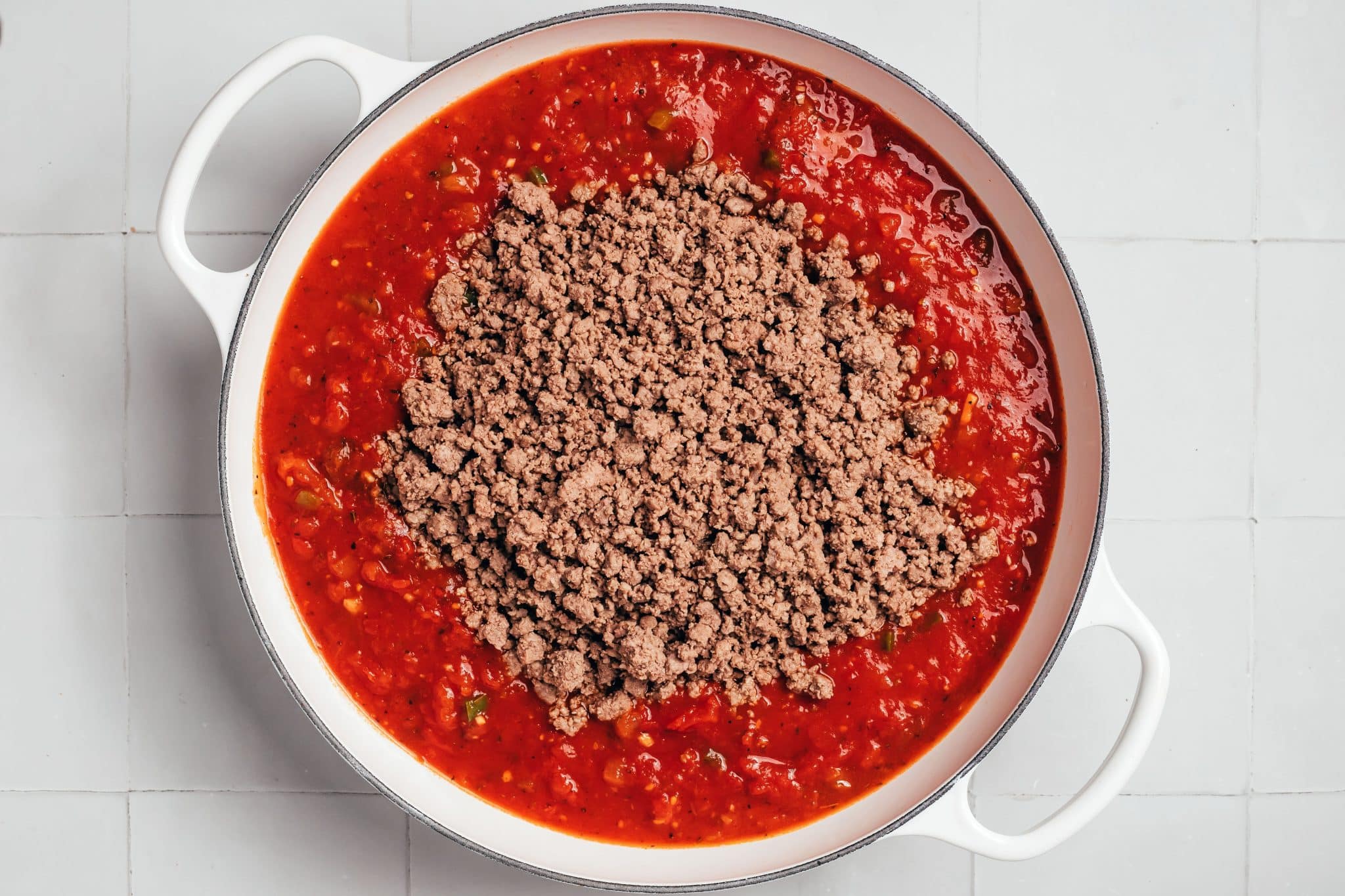 A generous mound of cooked, crumbled ground beef sits in the center of a white enameled Dutch oven filled with bright red tomato sauce. Small pieces of diced tomatoes, softened onions, green bell peppers, and dark flecks of seasoning are visible throughout the sauce surrounding the beef.