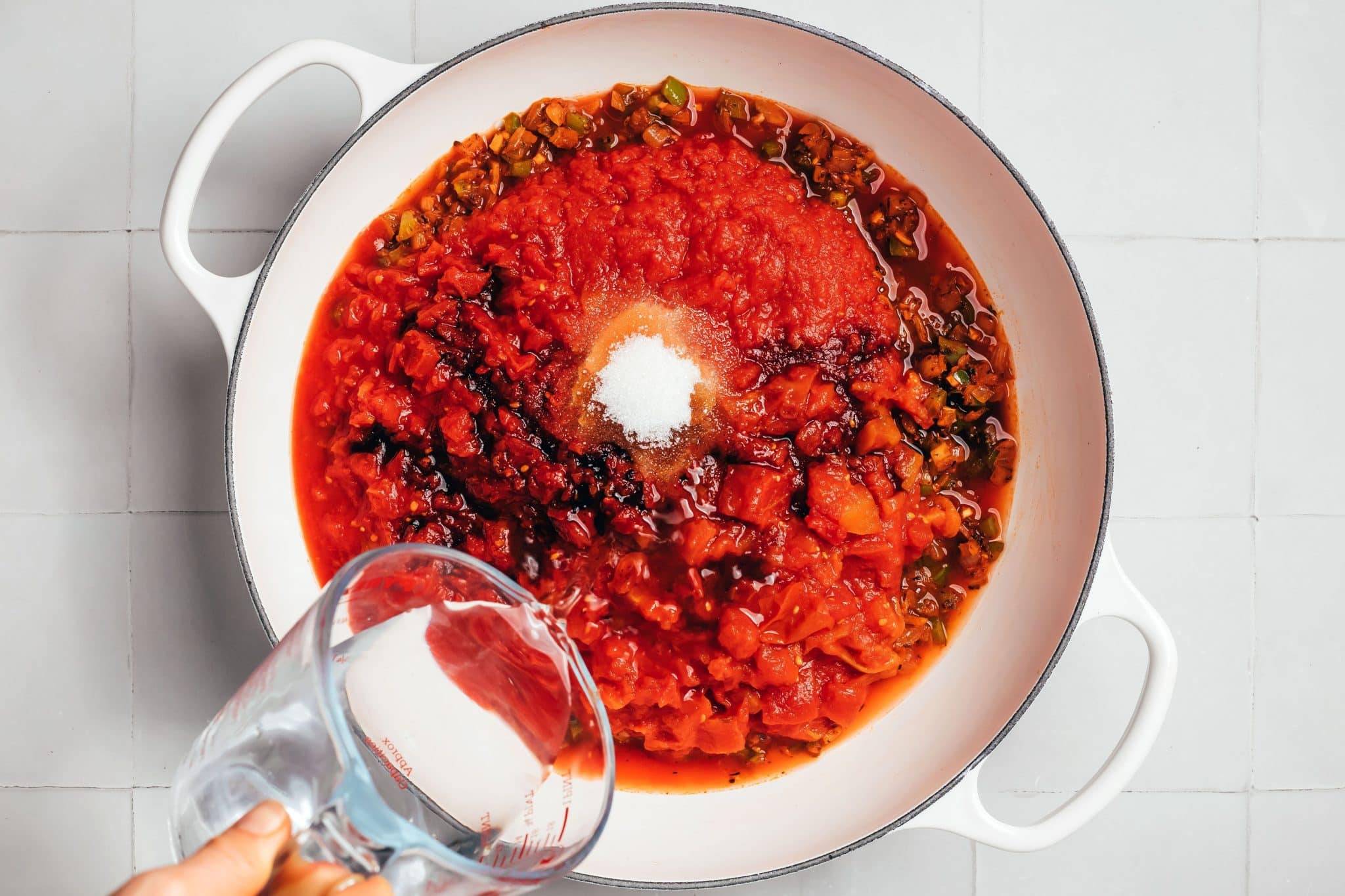 A white enameled Dutch oven filled with diced tomatoes, crushed tomatoes, and tomato sauce over sautéed onions and green bell peppers. A small mound of white sugar and dark streaks of Worcestershire sauce are visible among the red tomato mixture. A hand holds a tilted glass measuring cup to add water.