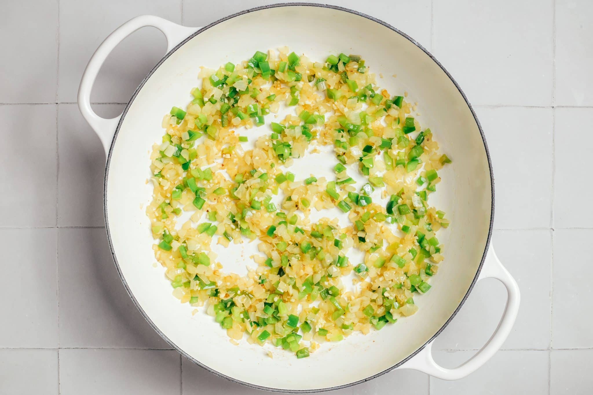 Finely diced green bell pepper mixed with softened yellow onions and small pieces of garlic in a white enameled Dutch oven. The onions are translucent and lightly golden, while the bell pepper pieces remain bright green and are beginning to soften.