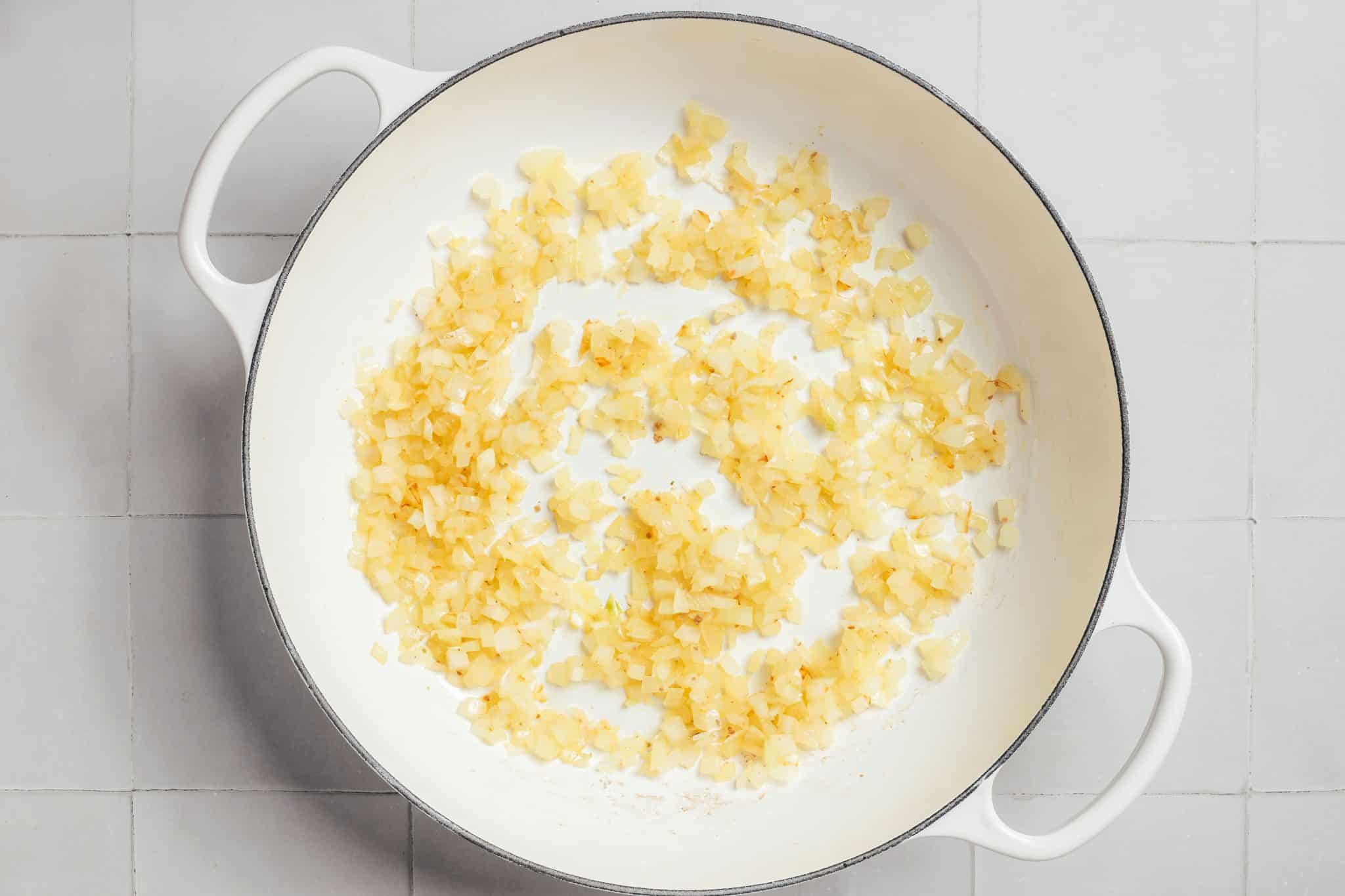 Finely diced yellow onions softened in a large white enameled Dutch oven. The onions are translucent and pale golden, with a few lightly browned edges. They are spread in a loose ring across the bottom of the pot, leaving much of the white cooking surface visible.