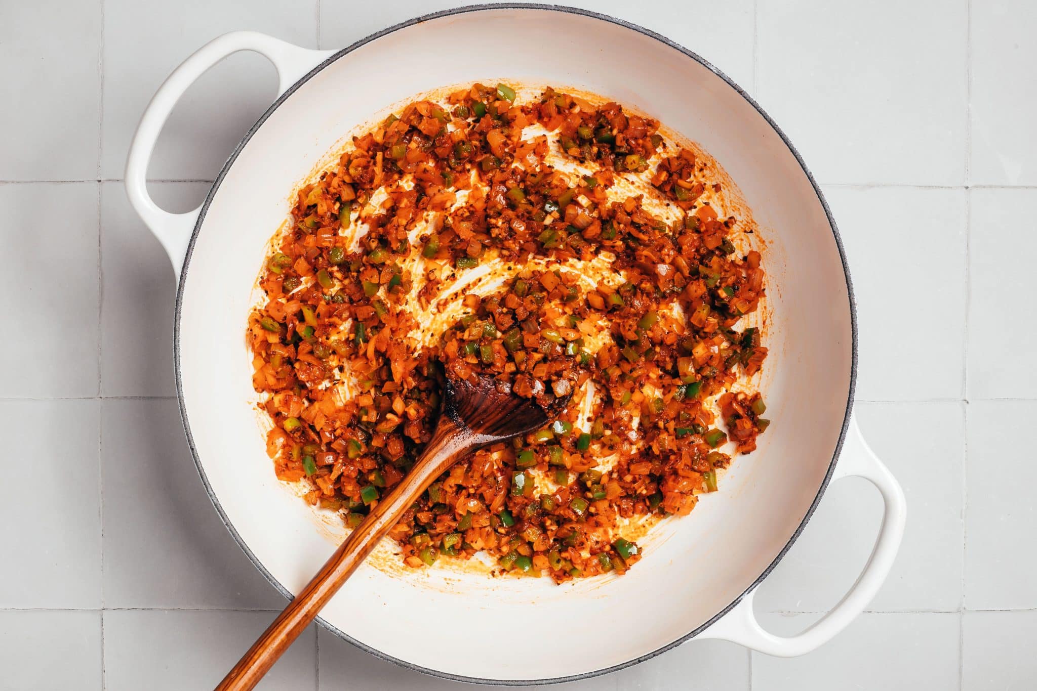 Finely diced onions and green bell peppers coated in a reddish-orange mixture of tomato paste and dried herbs inside a white enameled Dutch oven. Dark flecks of seasoning are visible throughout the vegetables, and a wooden cooking spoon rests in the center of the pot.