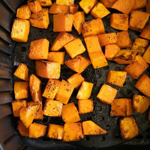 Cooked seasoned butternut squash cubes in the basket of a large air fryer.