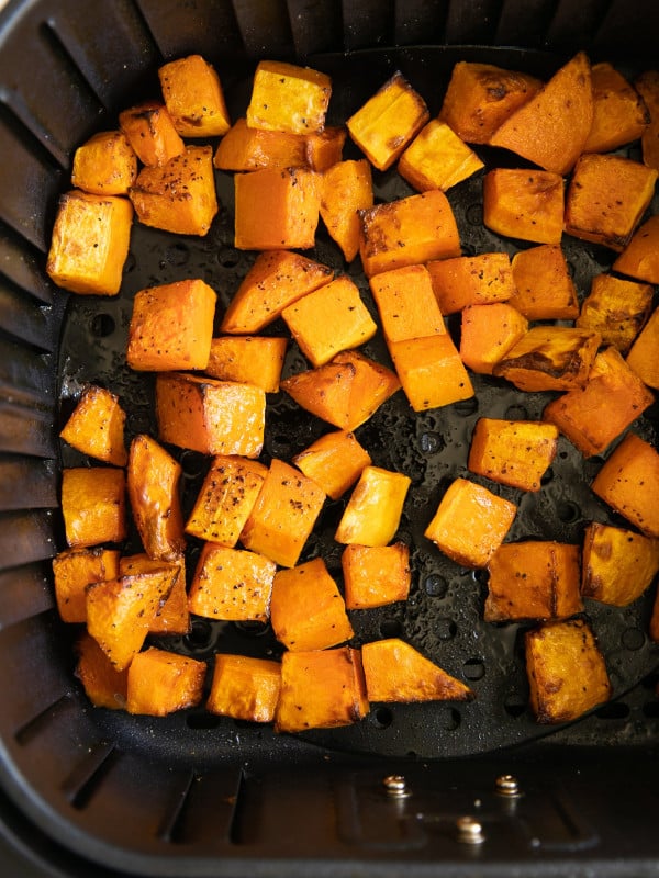 Cooked seasoned butternut squash cubes in the basket of a large air fryer.