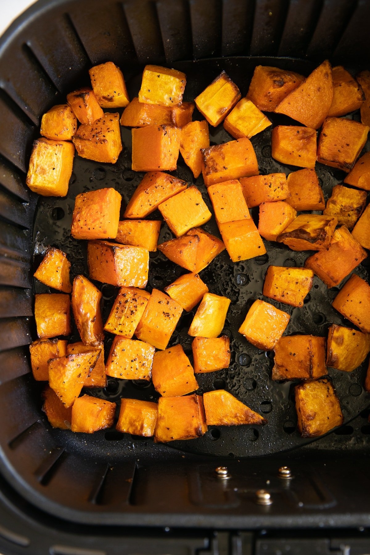 Cooked seasoned butternut squash cubes in the basket of a large air fryer.