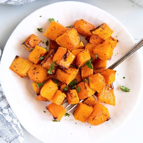 Small white plate filled with perfectly tender butternut squash cubes cooked in the air fryer with charred tips and garnished with fresh parsley.