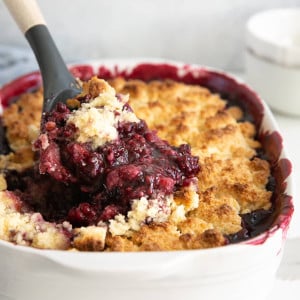 Spatula scooping hot blackberry cobbler from a white baking dish.
