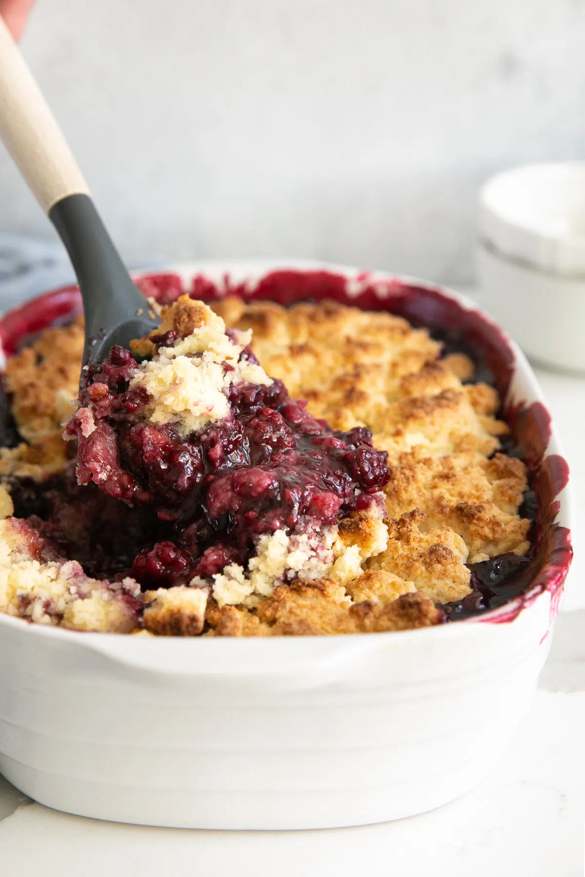 Spatula scooping hot blackberry cobbler from a white baking dish.