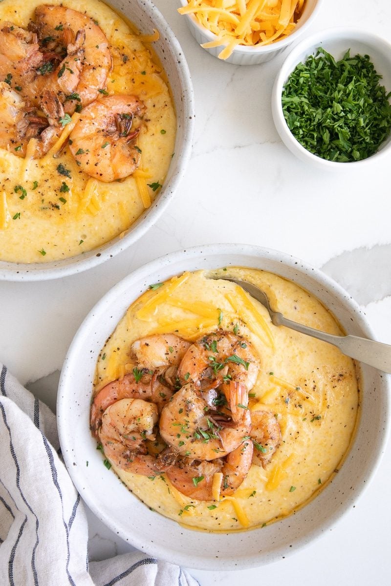 Two white bowls filled with cheesy grits topped with cooked shrimp and garnished with shreddd cheddar cheese and fresh parsley.