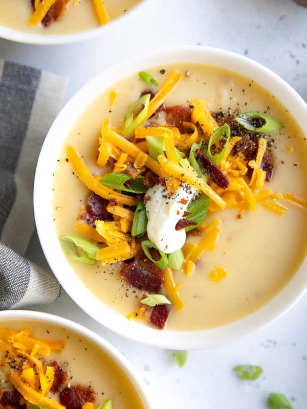 White bowl of creamy potato soup topped with shredded cheddar cheese, sliced green onions, bacon pieces, a dollop of sour cream, and black pepper on a light countertop, with other soup bowls partially visible in the background.