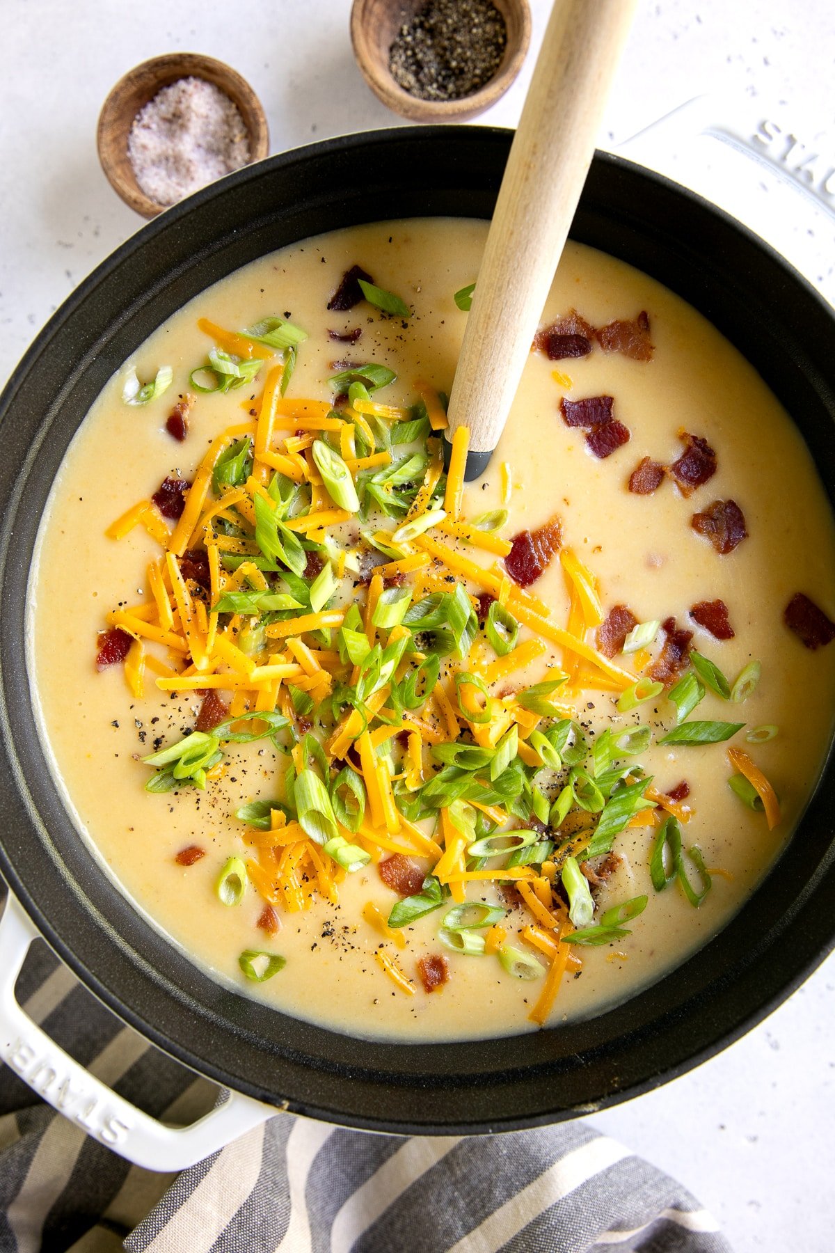 Overhead view of creamy potato soup in a black Dutch oven topped with shredded cheddar cheese, sliced green onions, bacon pieces, and black pepper, with a wooden spoon in the pot and small bowls of salt and pepper in the background.
