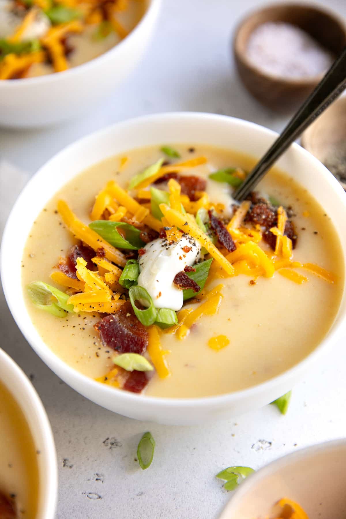 White bowl of creamy potato soup topped with shredded cheddar cheese, sliced green onions, bacon pieces, a dollop of sour cream, and black pepper on a light countertop, with other soup bowls partially visible in the background.