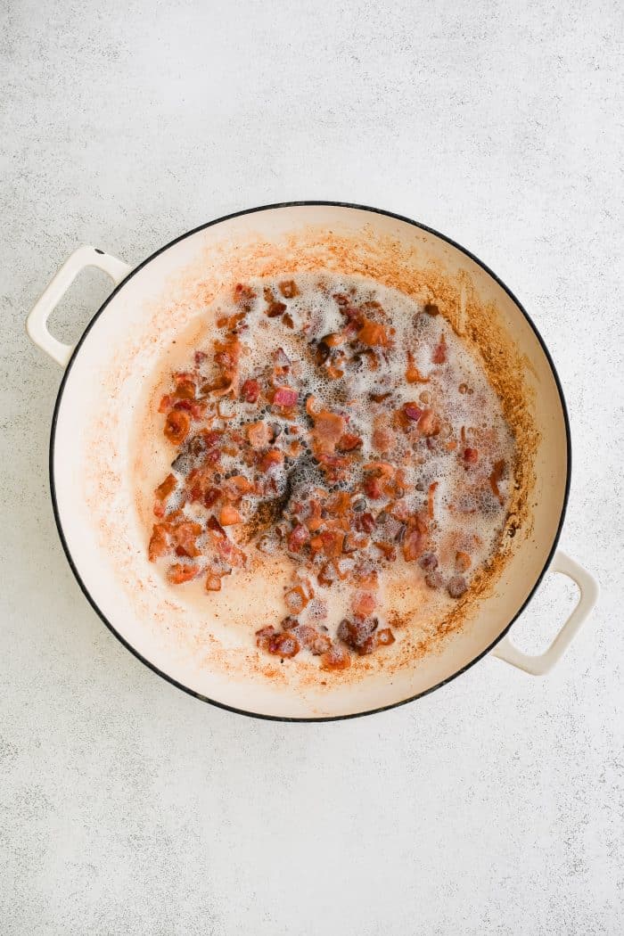 Overhead view of chopped bacon pieces cooking in a white enameled Dutch oven with rendered bacon fat and browned bits on the bottom of the pot, set on a light gray textured countertop.