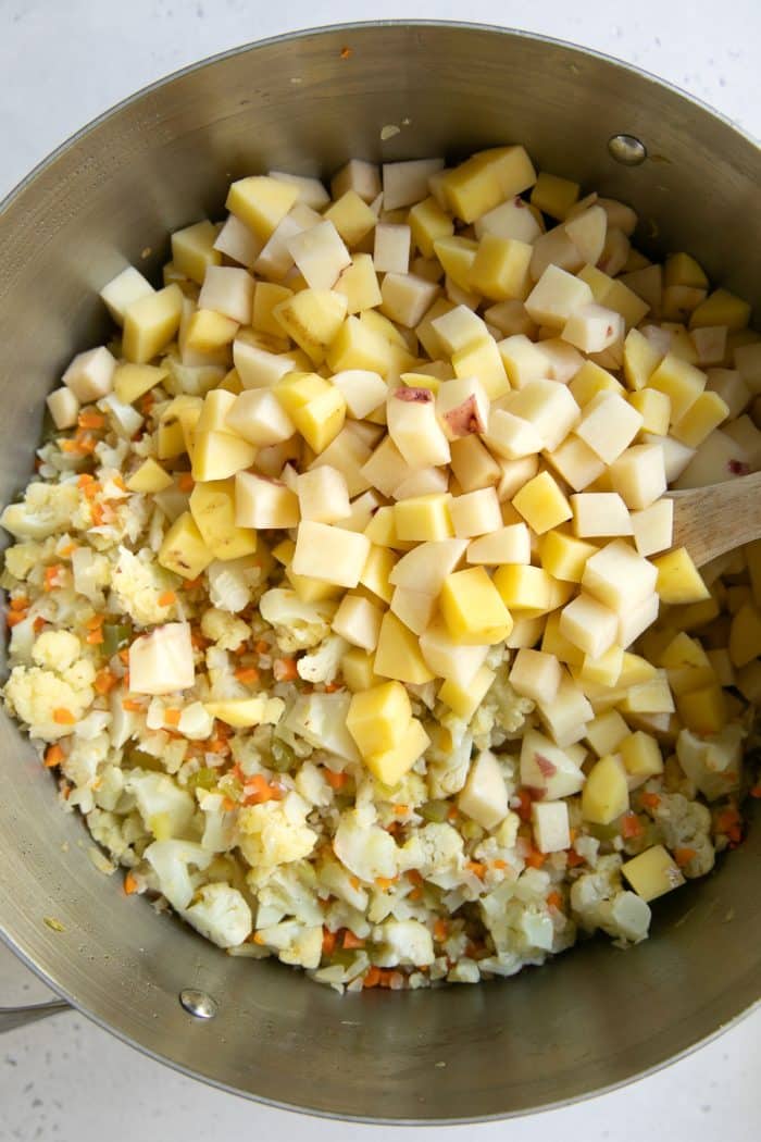 Diced potatoes added to a large stainless steel pot with cooked vegetables and cauliflower, with a wooden spoon resting along the right side.