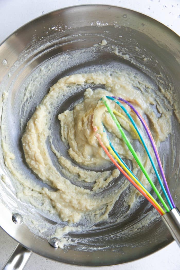 Butter and flour mixture forming a thick roux in a stainless steel saucepan, being whisked with a rainbow silicone whisk on a white countertop.
