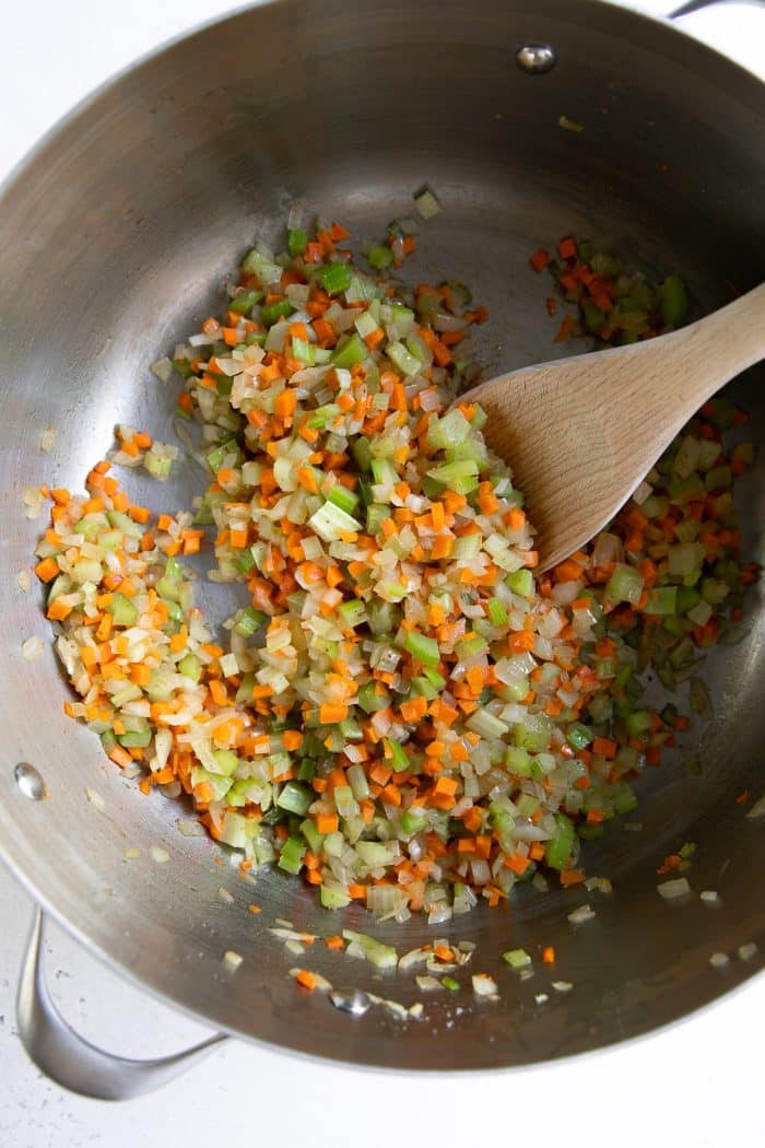 Chopped onions, carrots, and celery sautéing in a large stainless steel pot with a wooden spoon on the right side of the pot.