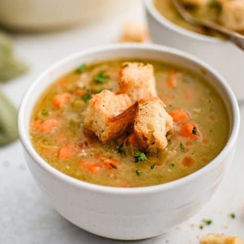 White soup bowl filled with split pea soup with chunks of ham and carrots and topped with homemade croutons and fresh parsley.