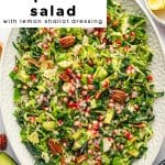 Vertical image of shaved Brussels sprout salad on a white platter with pomegranate, avocado, and pecans, styled with lemons and dressing bowls