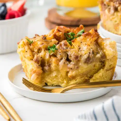 Serving of breakfast casserole made with bread, sausage, egg, and cheese on a white plate with a gold fork, orange juice, and a small bowl filled with fruit in the background.