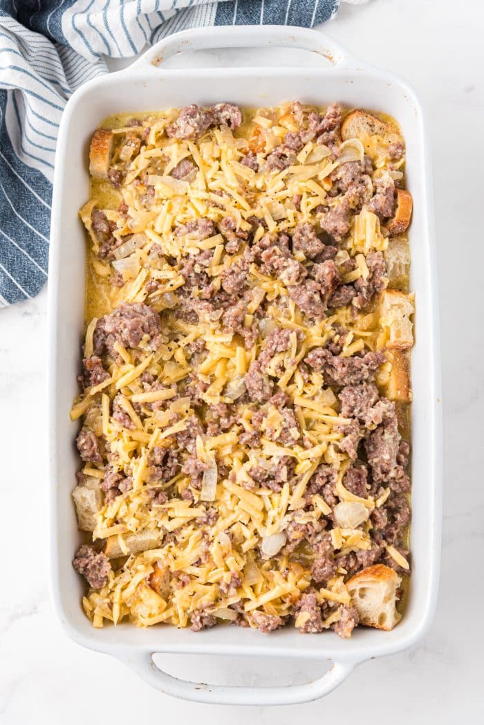 Large white casserole dish filled with a bottom layer of day-old bread and drizzled with sausage, egg, milk, and cheese mixture over the top.