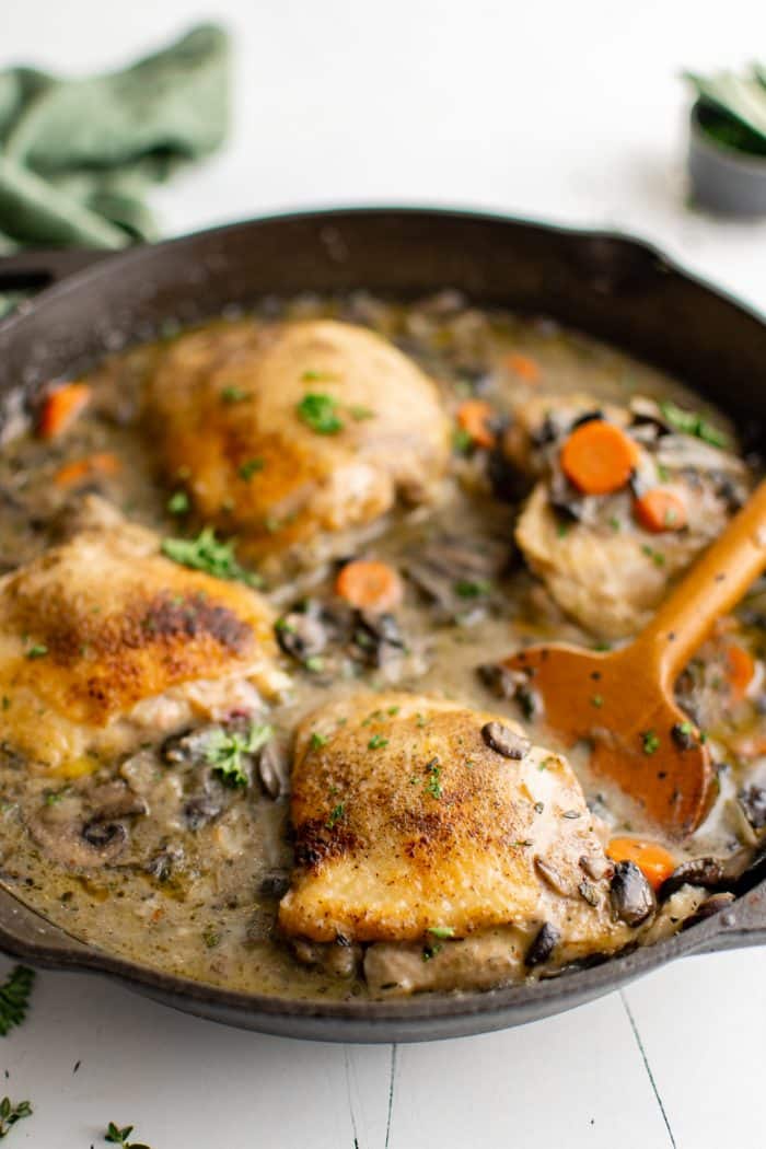 Large cast iron skillet filled with four large browned chicken thighs braised in a herb and white wine sauce filled with carrots, onions, and mushrooms.