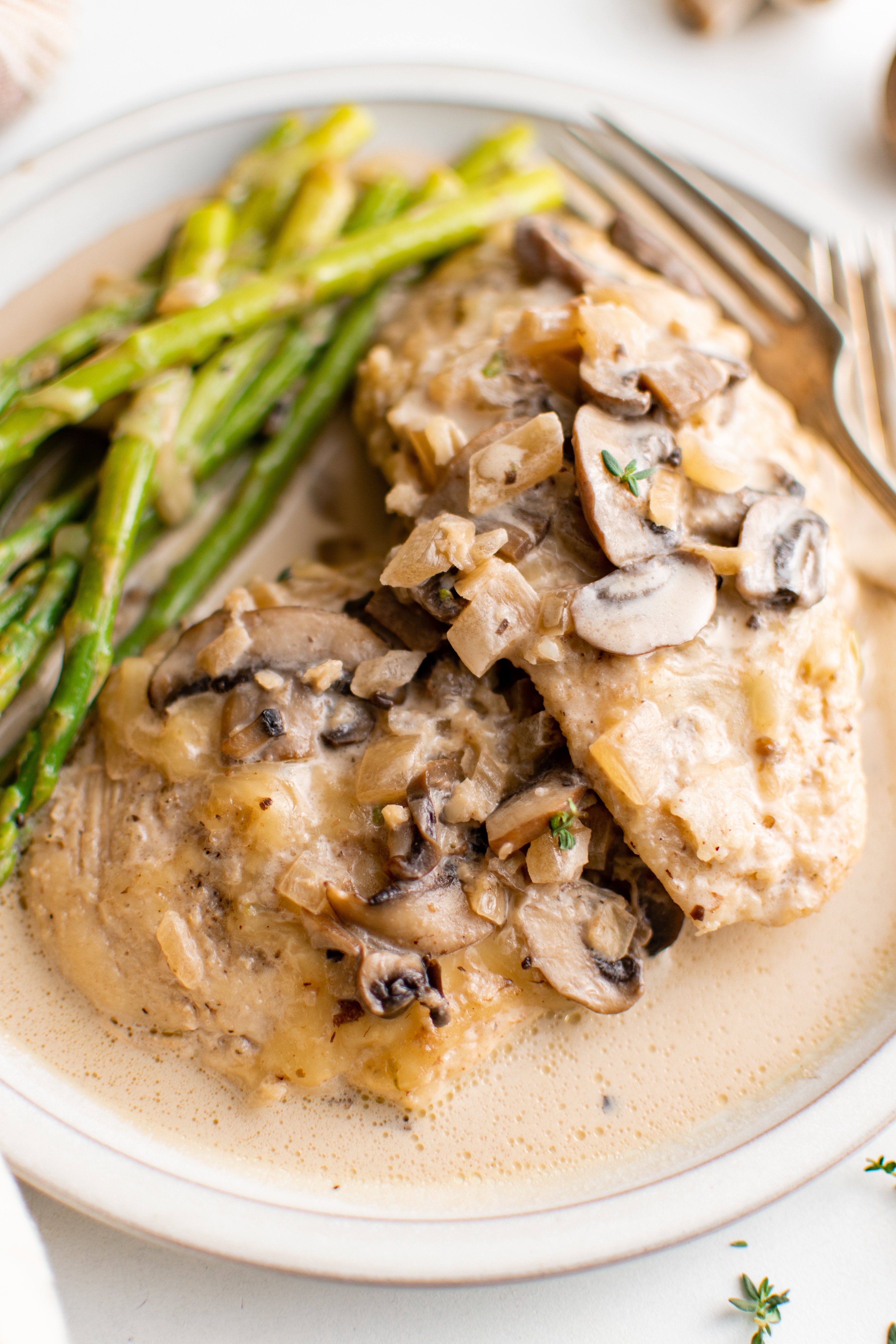 White dinner plate with creamy mashed potatoes topped with chicken madeira and mushrooms and a creamy madeira sauce with a side of asparagus.