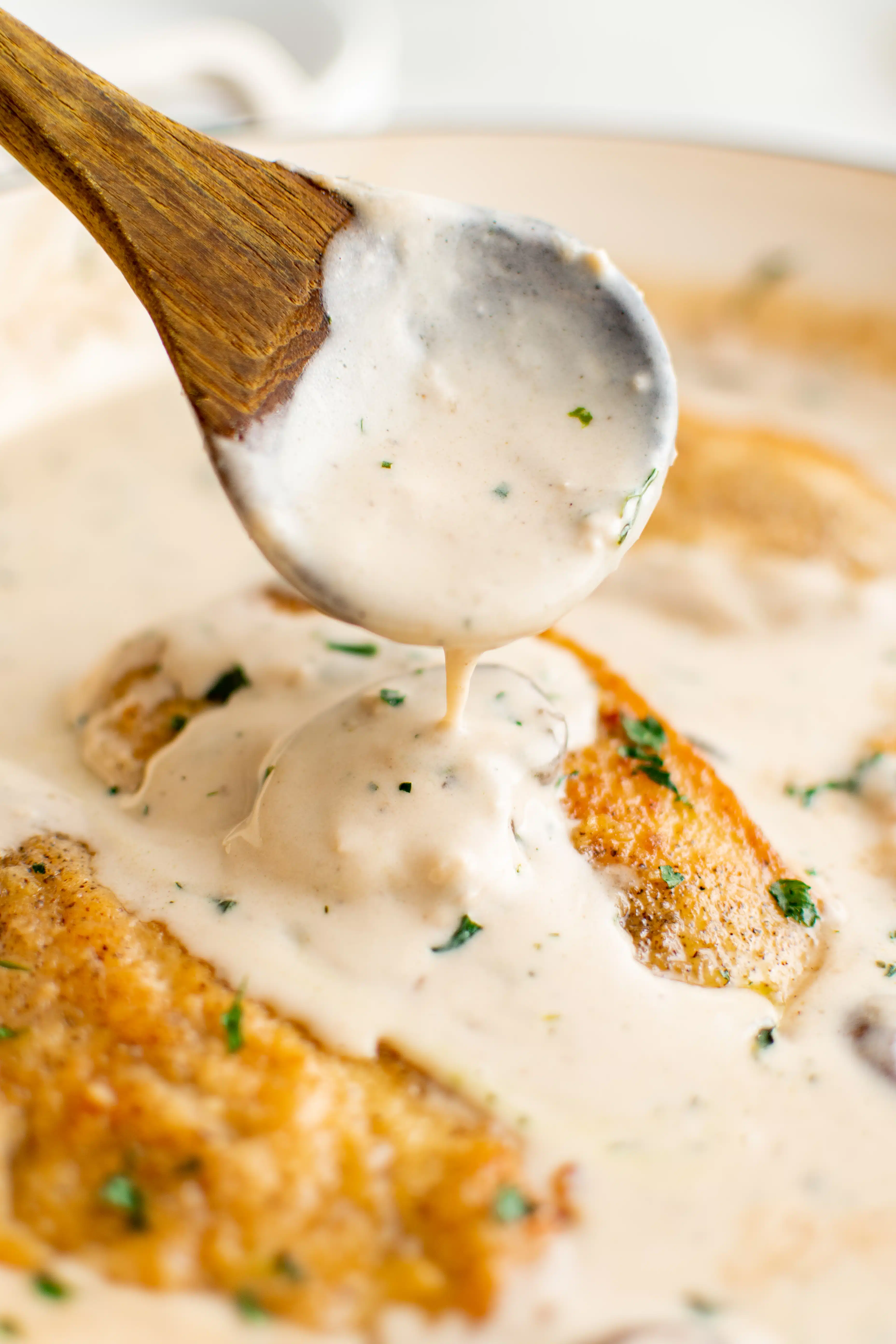 Wood serving spoon pouring homemade marsala cream sauce over a cooked chicken cutlet in a large pan filled with marsala cream sauce.