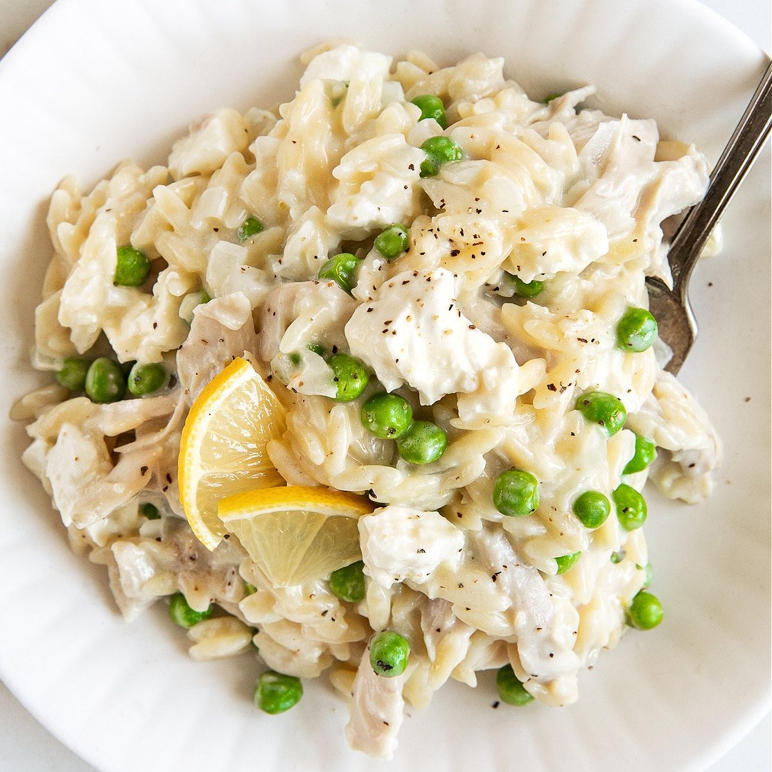 White plate filled with cooked orzo in a lemon cream sauce with shredded chicken, peas, and feta cheese.