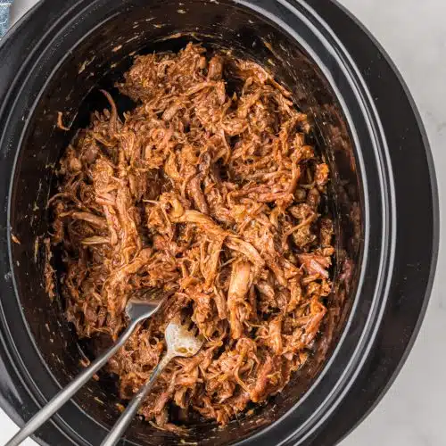 Shredded cooked pork shoulder mixed with bbq sauce in a large slow cooker.